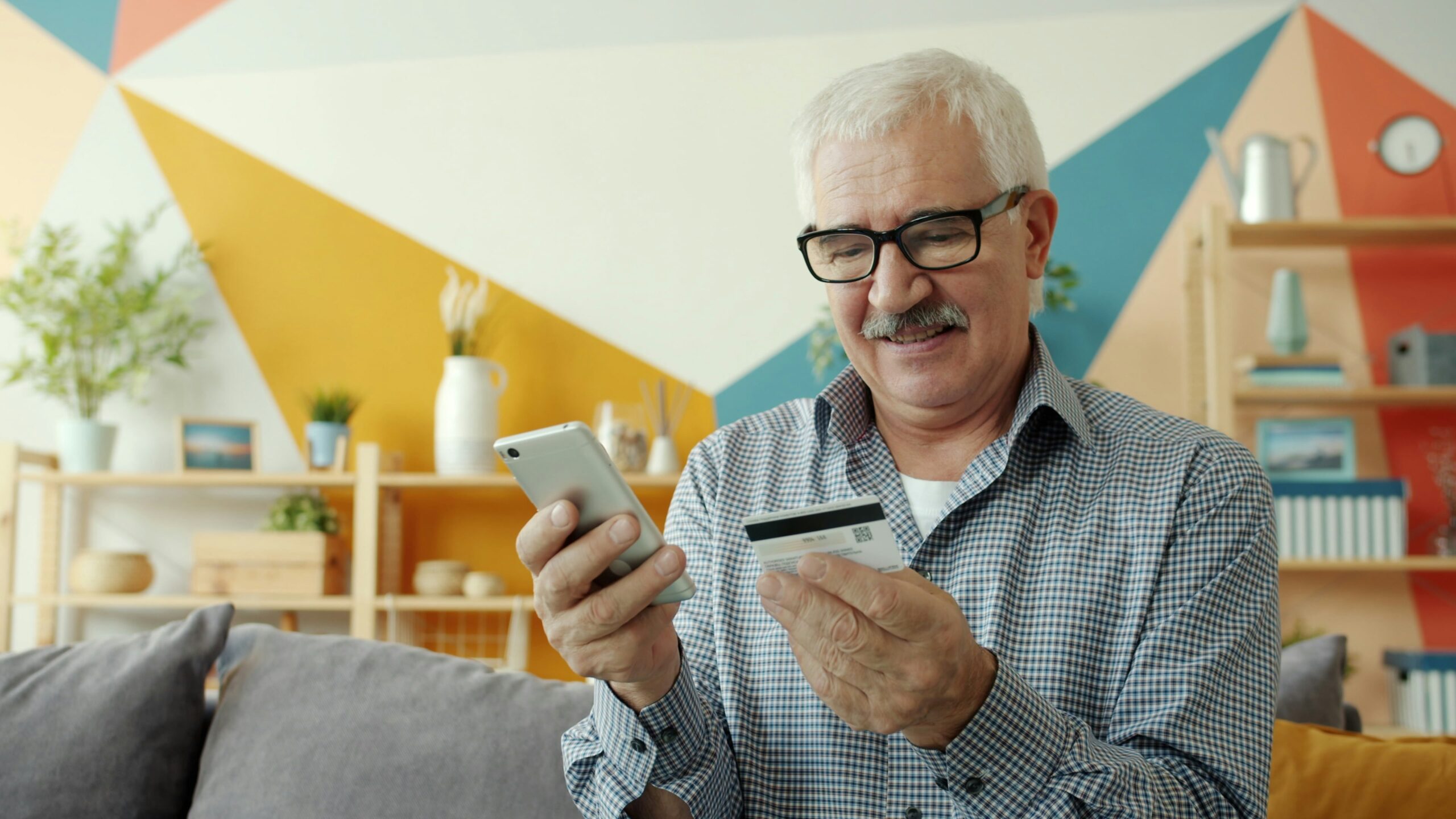 Good-looking retired man is shopping online using smartphone paying with plastic card from apartment enjoying modern technology and electronic money.